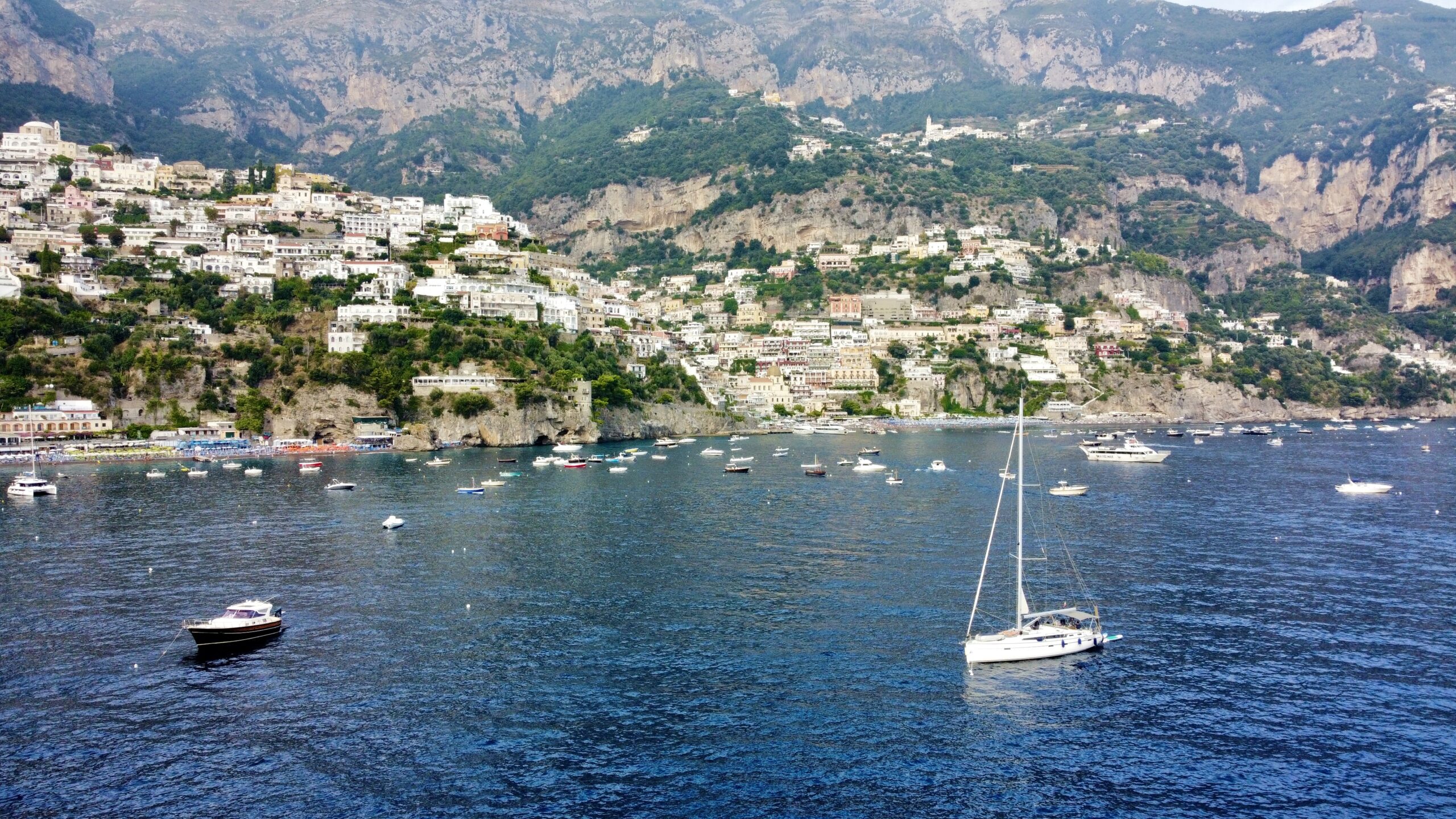 Aerial view of a yacht sailing in the turquoise waters of the Amalfi Coast, surrounded by lush green landscapes and clear skies, captured by a drone.